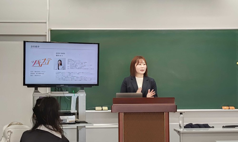 桜のどかが大学の講義に登壇し、ツギステの会社紹介スライドを前に元アイドルのセカンドキャリアについて話している様子