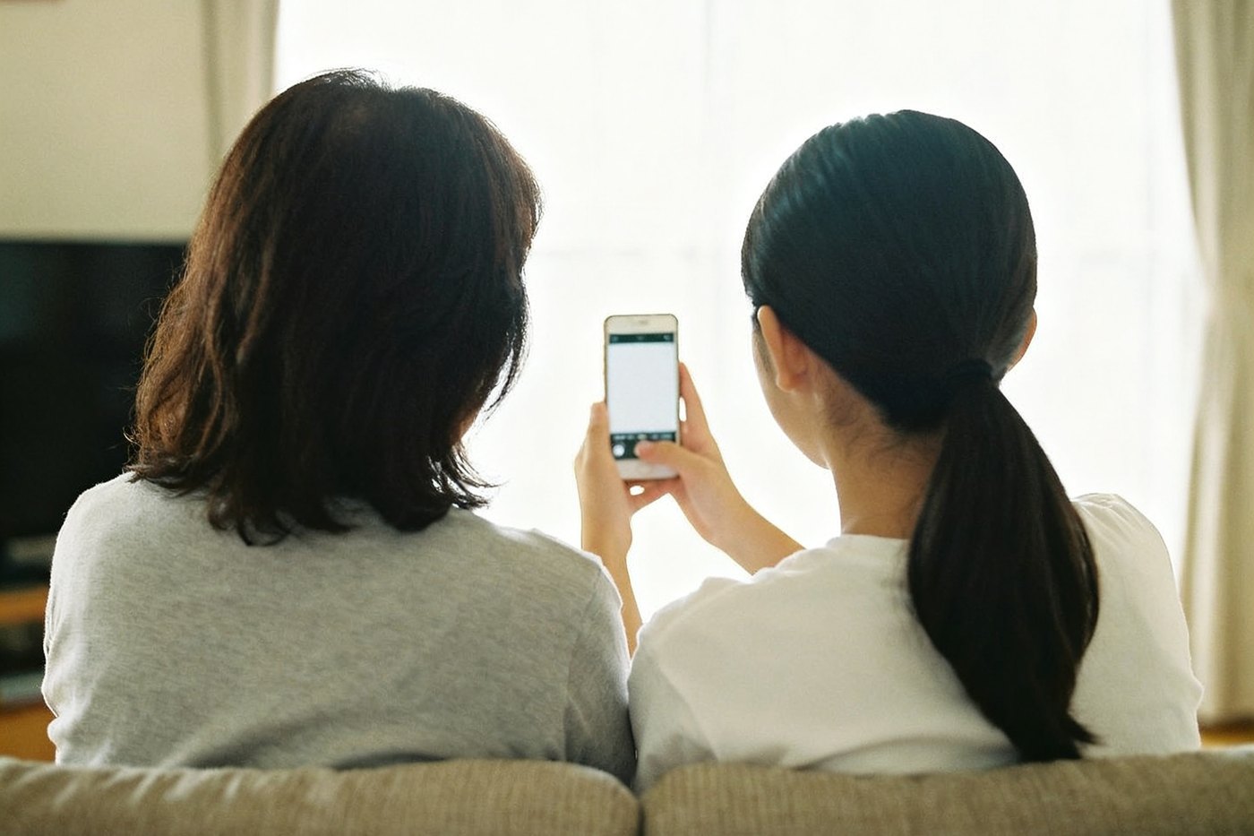 ソファに並んで座る親子の後ろ姿。娘が持つスマートフォンを、二人で一緒にのぞき込んでいる
